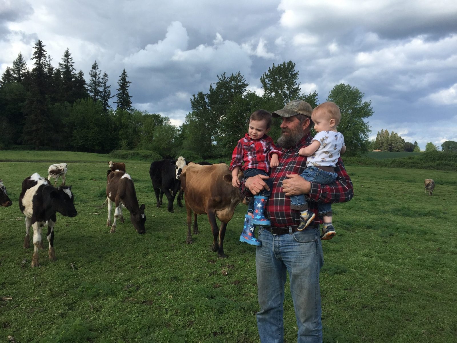A Hess family farmer holding two young children in the pasture as the dairy herd grazes behind them